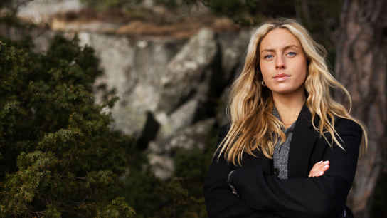 Kvinna med ljust hår och svart kavaj framför en skog